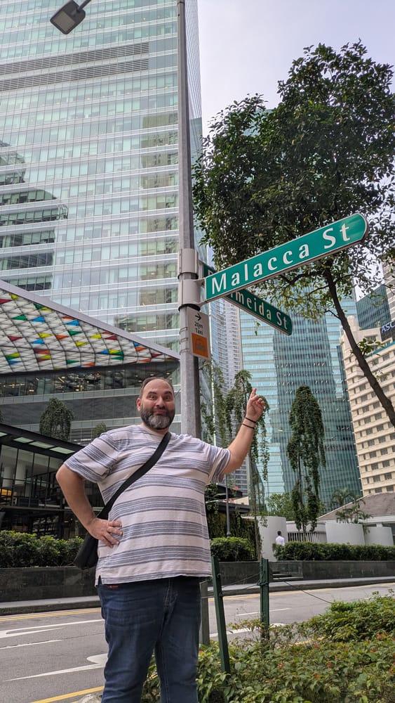 Me standing under the Malacca Street sign in Singapore's financial district, pointing up at it with a delighted expression