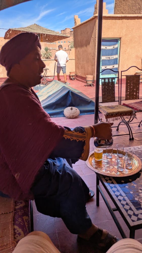 A man in a headwrap pouring mint tea into small glasses on a rooftop terrace in Morocco, with kasbah walls behind