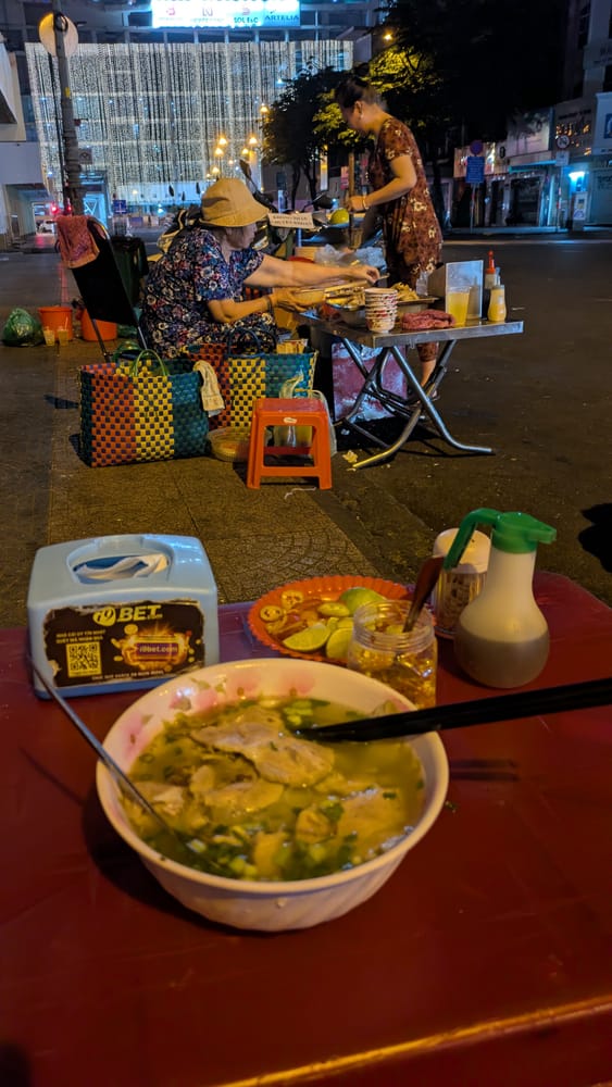 A bowl of noodle soup on a red plastic table at a night-time street stall in Ho Chi Minh City, with the two women running the stall working behind it